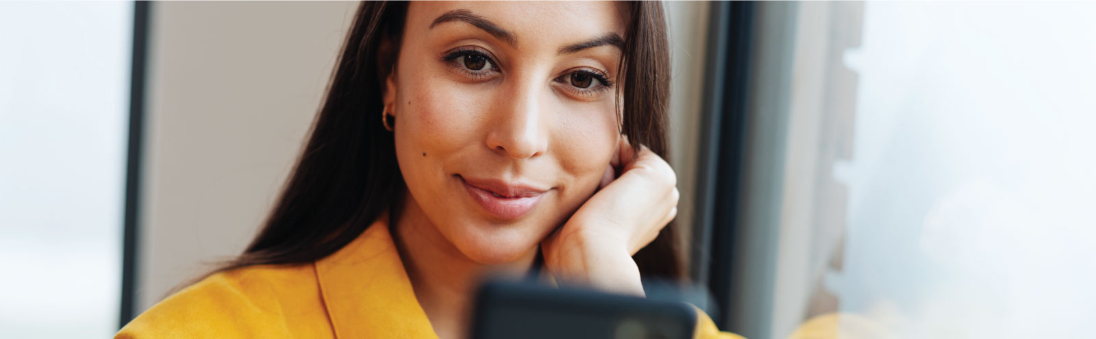 Woman in gold shirt leaning on a window looking at her cell phone