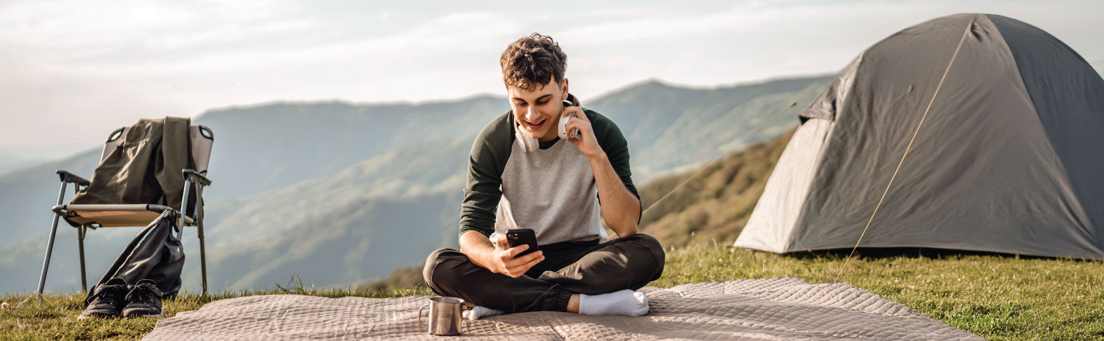 Young man holding his phone sitting on a blanket outside a tent in the mountains.